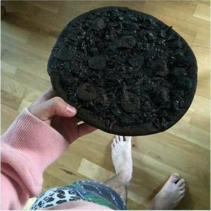 A hand holding a completely burnt, black pizza over a wooden floor, a total kitchen fail.