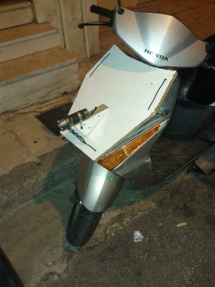 A DIY fail: a flashlight duct-taped to a white board acting as a makeshift headlight on a Honda scooter.