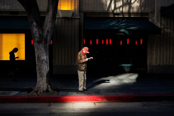 Perfectly timed street photo by Jeremy Paige of a man reading a book under a spotlight at night.