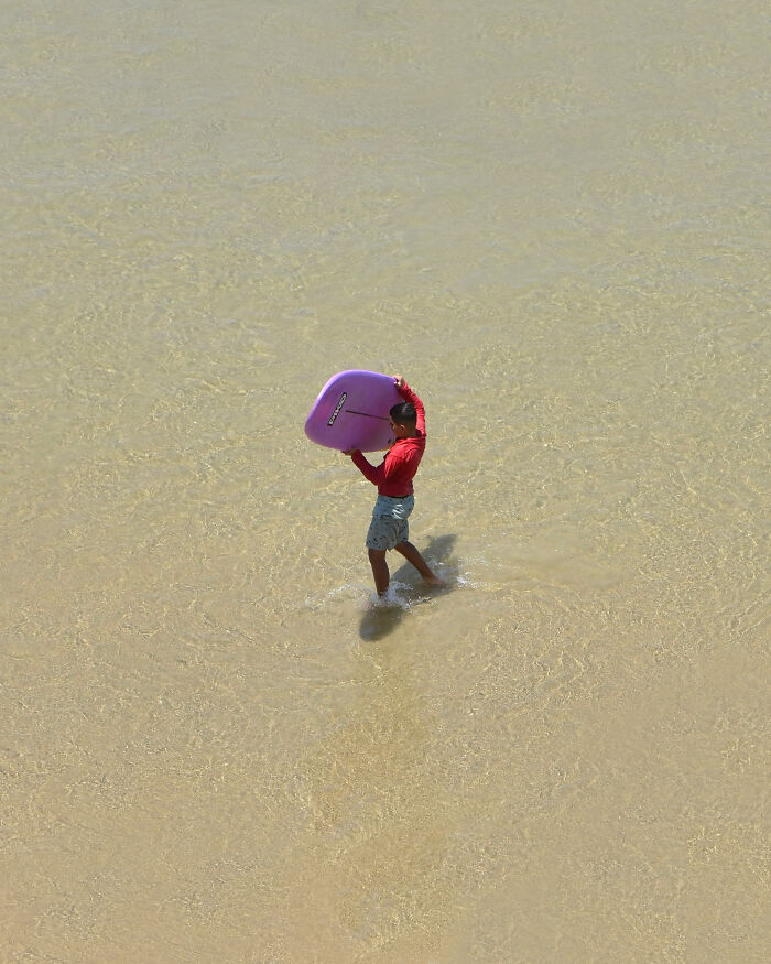 A photographer uses negative space to capture a person carrying a purple surfboard in shallow beach water.