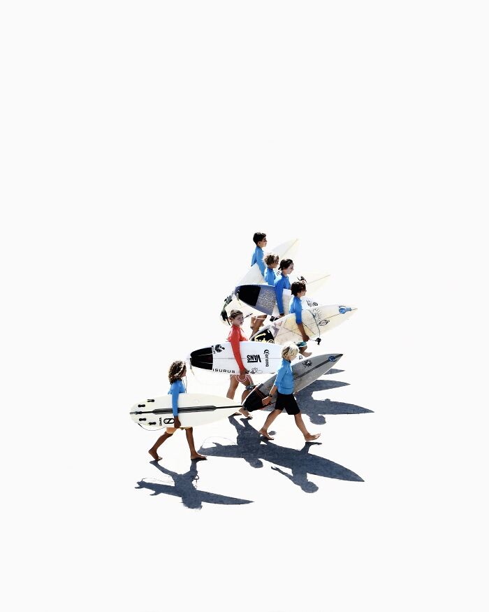 A line of young surfers walks with their boards across a stark white background, creating minimalist art using negative space.