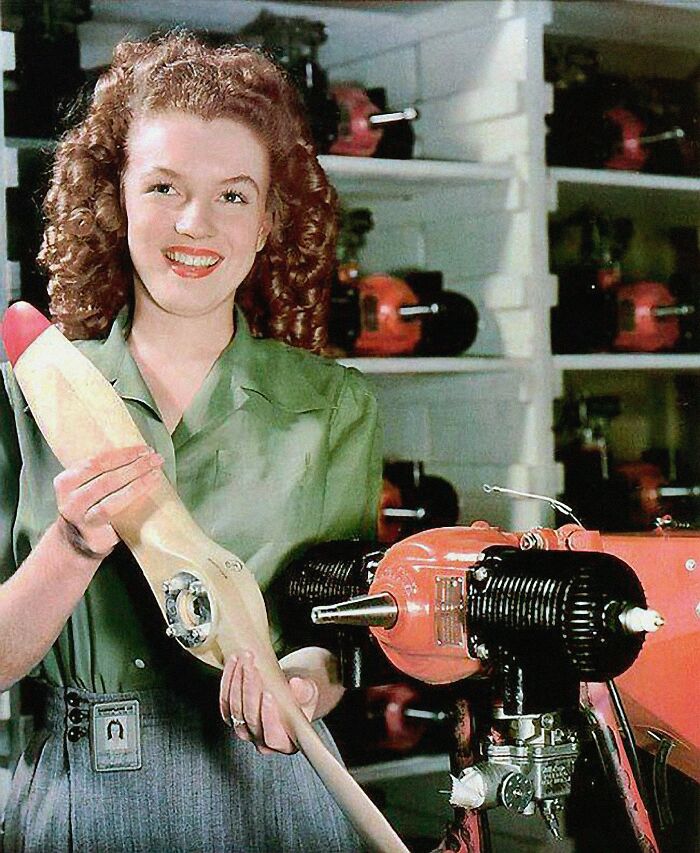 Marilyn Monroe, a prominent woman in history, smiles holding a propeller, with machinery visible in the background.
