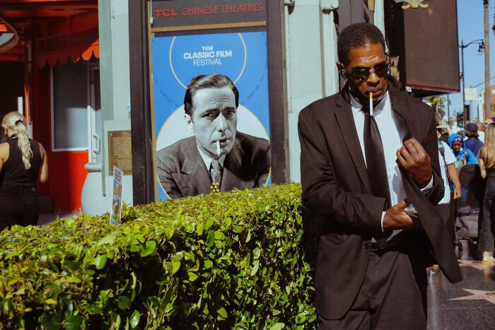 A man in a suit smoking, perfectly timed street photo with a classic film festival poster in the background by Jeremy Paige.