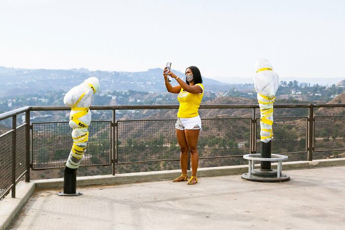 Perfectly timed street photo by Jeremy Paige of a woman in a yellow shirt taking a photo of wrapped sculptures.