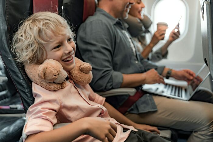A happy young boy wearing a neck pillow on an airline seat, smiling and looking out.