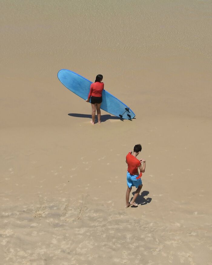 A photographer uses negative space to capture two people with a surfboard on a minimalist beach.