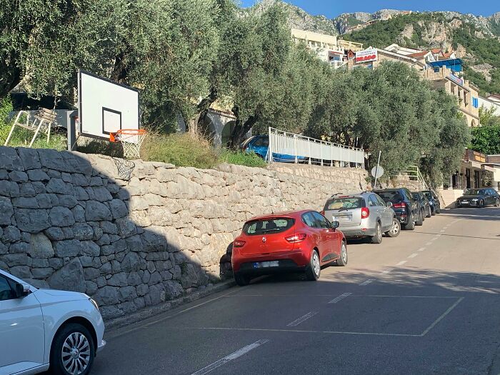 A basketball hoop positioned awkwardly above a steep stone wall, next to parked cars, a playground fail.