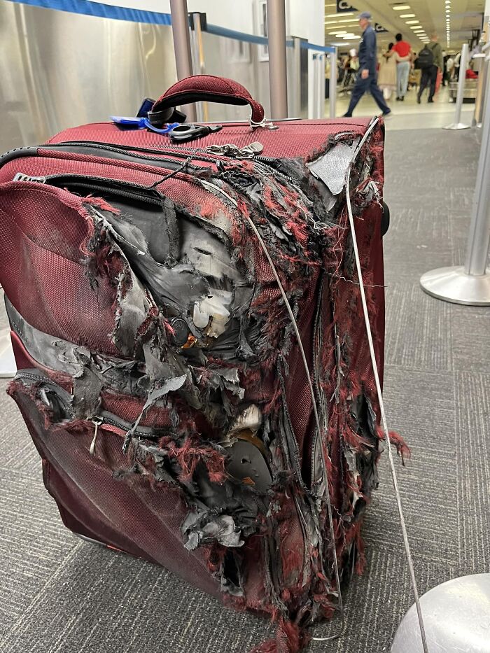 A severely damaged red suitcase on an airport floor, showing a reality check for entitled people.