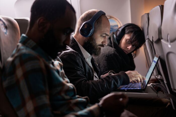 Passengers on an airplane with one man using a laptop and wearing headphones, illustrating entitled people getting a reality check.
