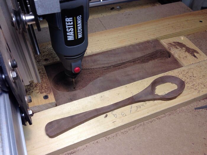 A partially carved wooden spoon with a large hole in the bowl, next to a CNC router, a clear DIY fail.
