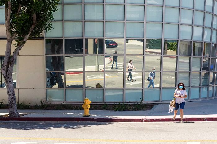 Perfectly timed street photo by Jeremy Paige capturing a woman walking past a reflective building showing street life.