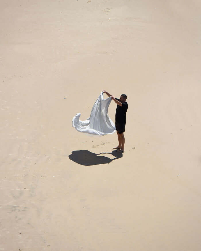 A person stands on a vast beach holding a white sheet, captured as minimalist art using negative space.