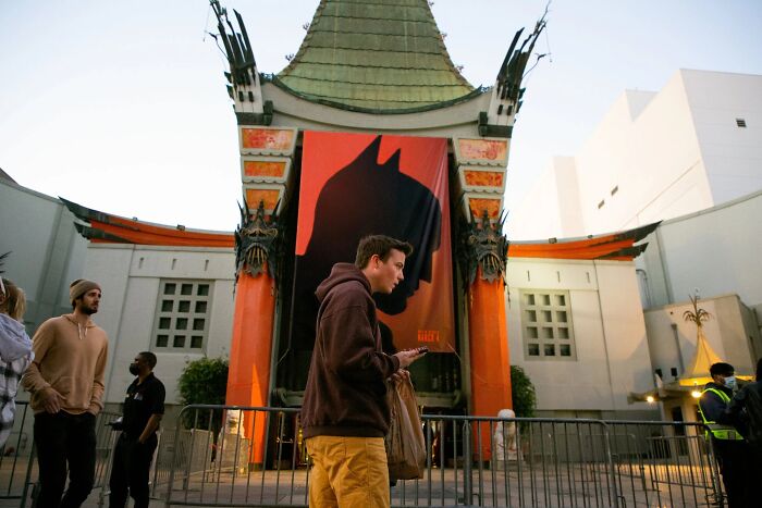A person standing in front of TCL Chinese Theatres with a Batman banner, a perfectly timed street photo by Jeremy Paige.