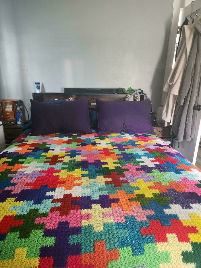Colorful crochet blanket with cross patterns covering a bed with purple pillows