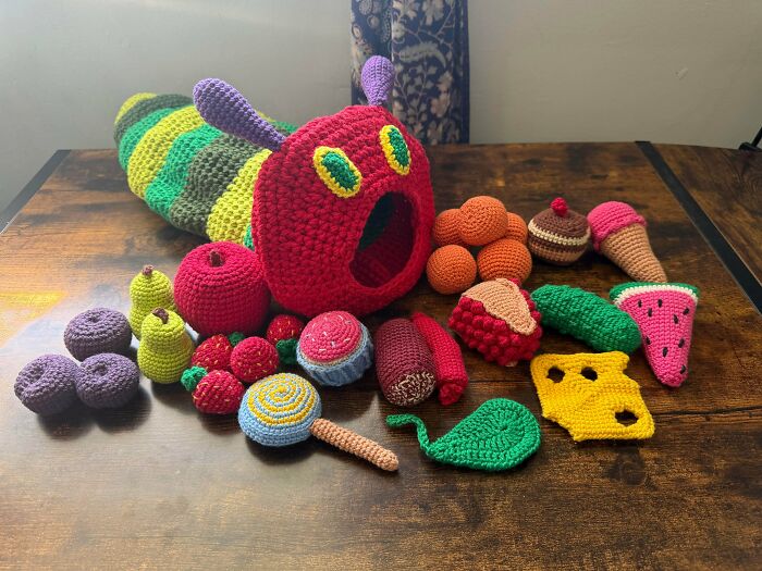 Collection of crocheted food items including a worm, fruits, and desserts on a wooden table