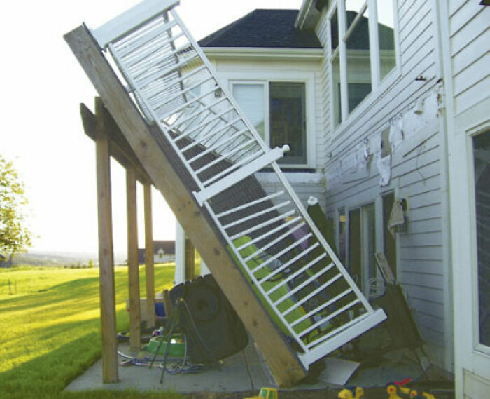 A DIY fail: a porch railing has completely detached and fallen over, leaning against the side of a house.