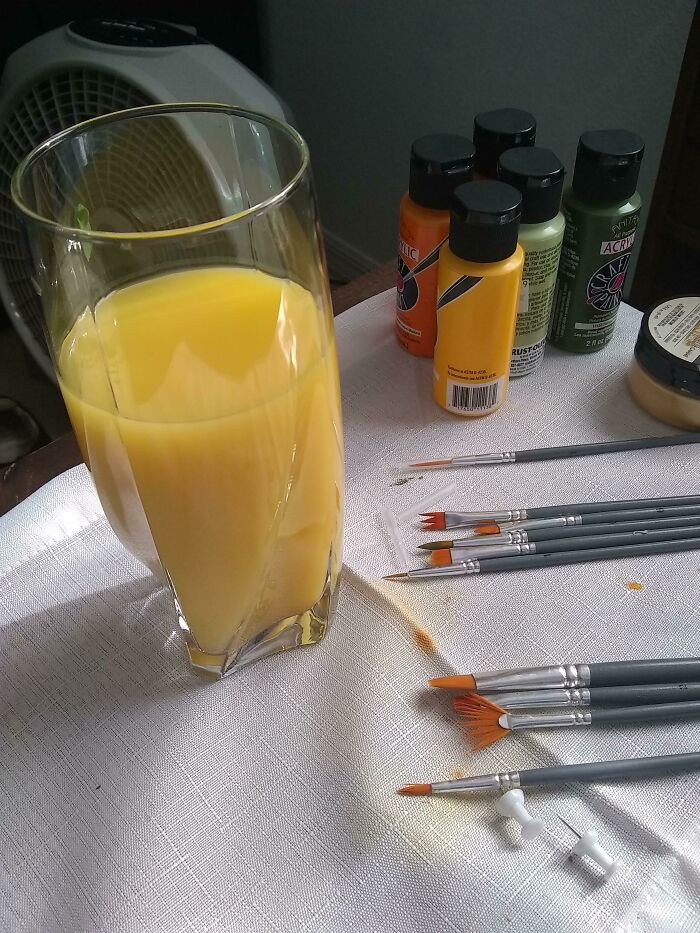 A glass of orange juice next to paint bottles and paintbrushes, resembling a forbidden food or drink.