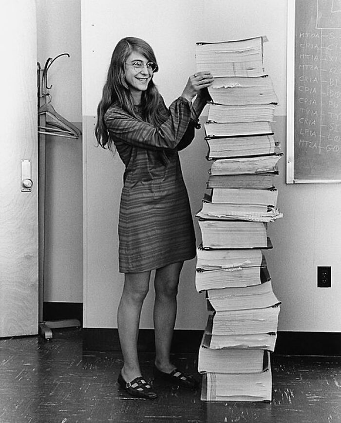 Margaret Hamilton, a leading woman in history, stands beside a tall stack of computer code, showcasing her work.