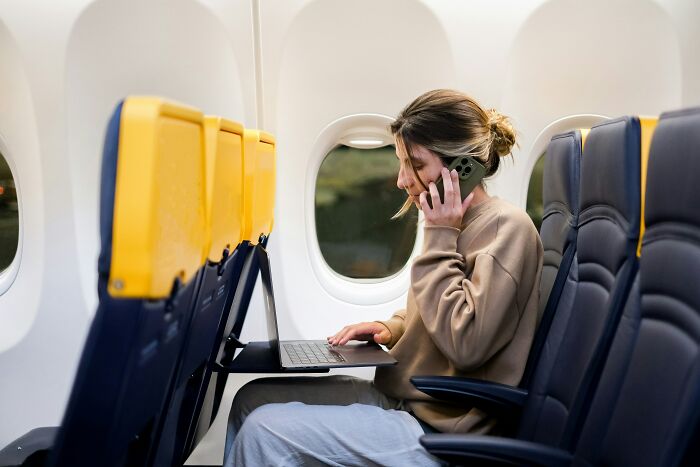 A woman talks on her phone and uses a laptop in an airplane, illustrating things cabin crew deal with.