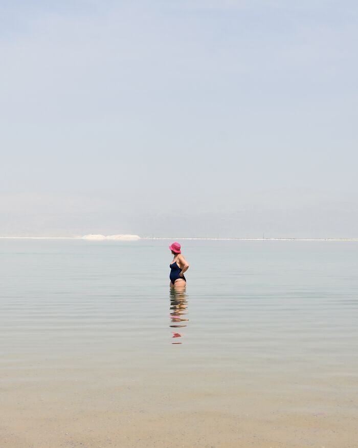 A person in a pink hat and dark swimsuit stands in calm, shallow water, exhibiting minimalist art of a beach scene with negative space.