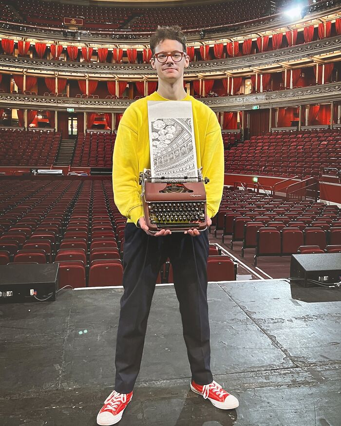 A man holds his typewriter art of a grand hall, standing on a stage with many red seats in the background.