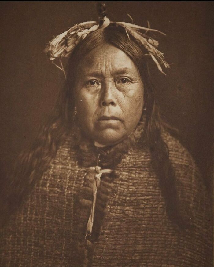 A Native American woman in history, captured in a solemn sepia-toned portrait wearing a headdress and woven garment.
