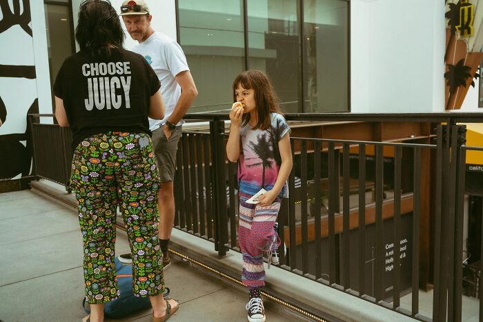 Perfectly timed street photo by Jeremy Paige of a girl eating a snack with adults nearby.
