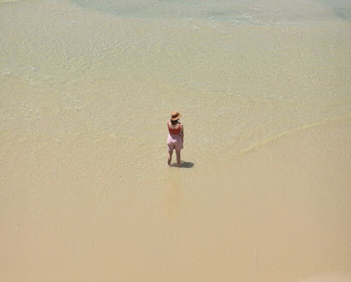 A person in a hat stands in clear, shallow water at the edge of a sandy beach, capturing minimalist art with negative space.