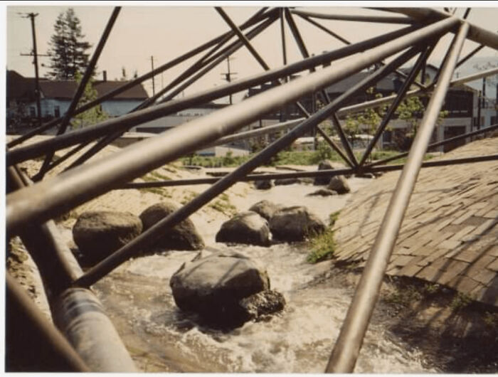 A narrow stream with rocks flowing under a metal playground structure, highlighting playground fails.