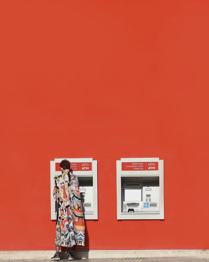 Photographer uses negative space: a person in a colorful coat at two ATMs against a vibrant red wall.