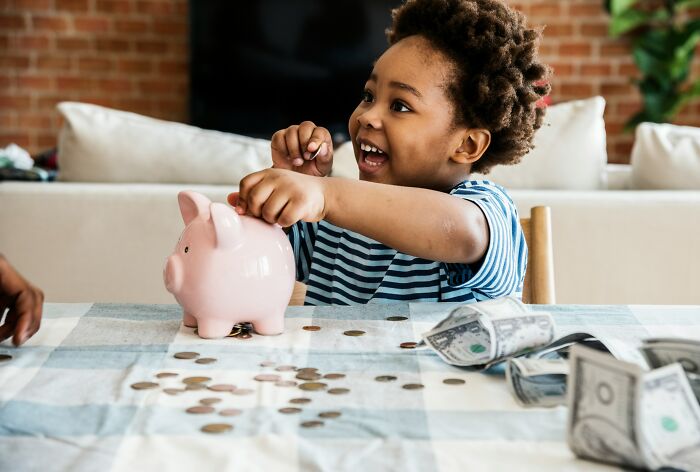 A happy young child putting coins into a pink piggy bank on a table with money.