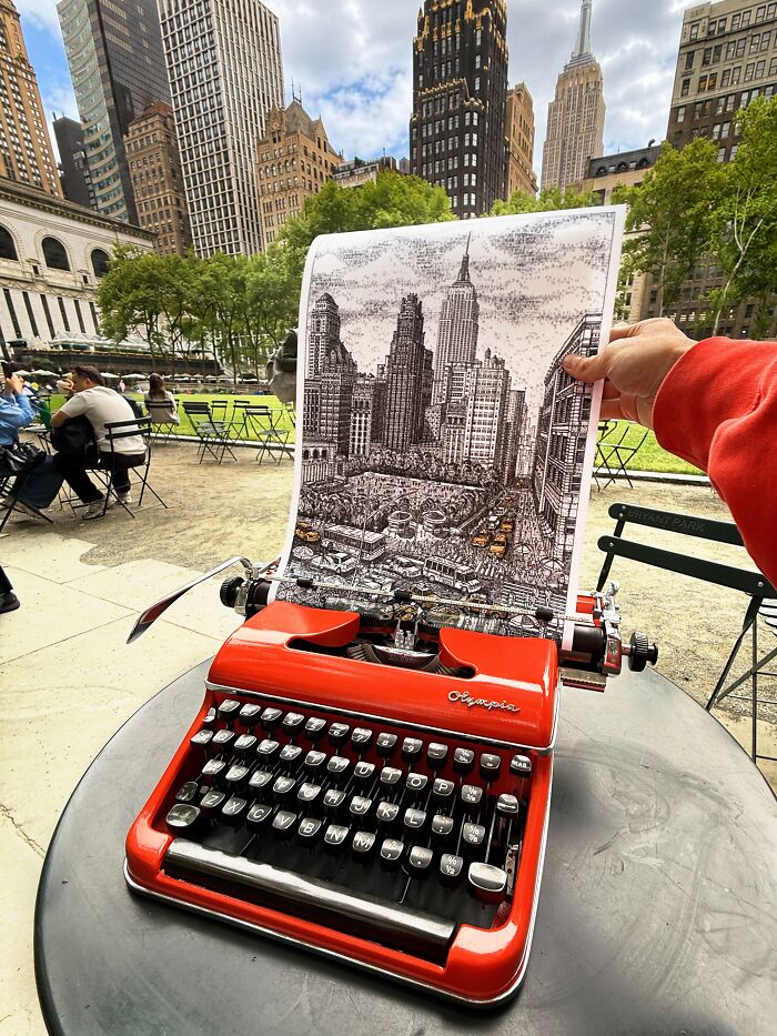 A red vintage typewriter in an outdoor setting, with a piece of detailed cityscape typewriter art being held above it.