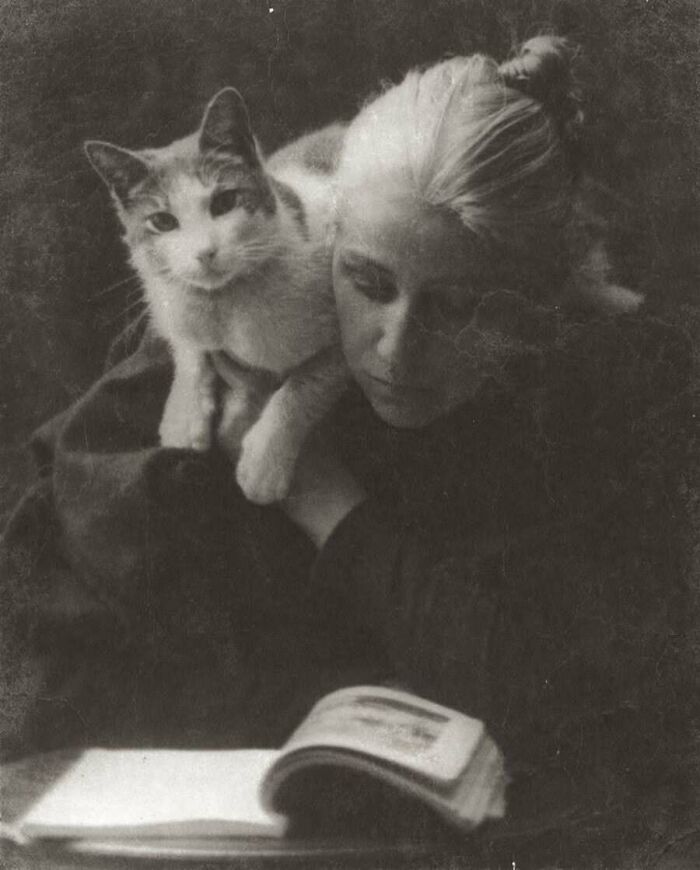 A black and white vintage photo of a woman holding a cat while reading a book