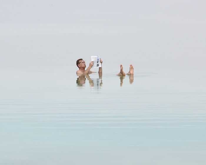 A man floats in serene water, reading a book, demonstrating minimalist art in a beach scene using negative space.