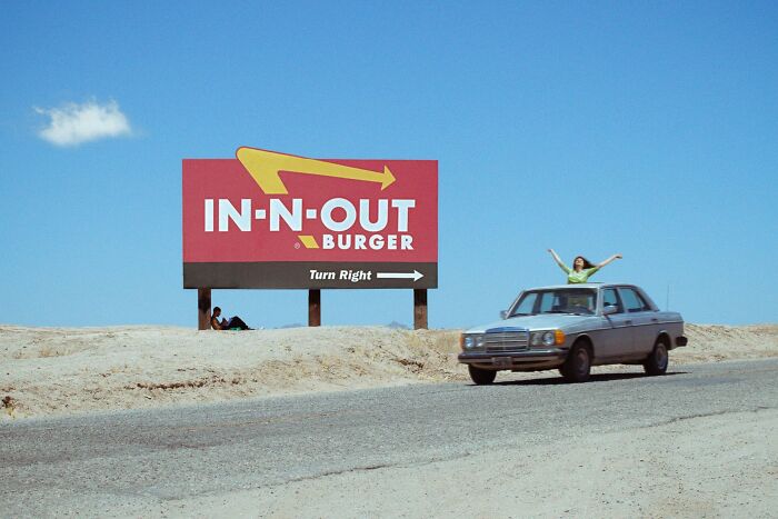 Perfectly timed street photo by Jeremy Paige of a person standing on a car roof next to an In-N-Out Burger sign.