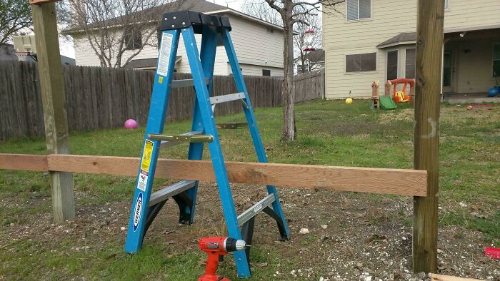 A DIY fail featuring a blue ladder holding up a wooden beam between two posts in a backyard.
