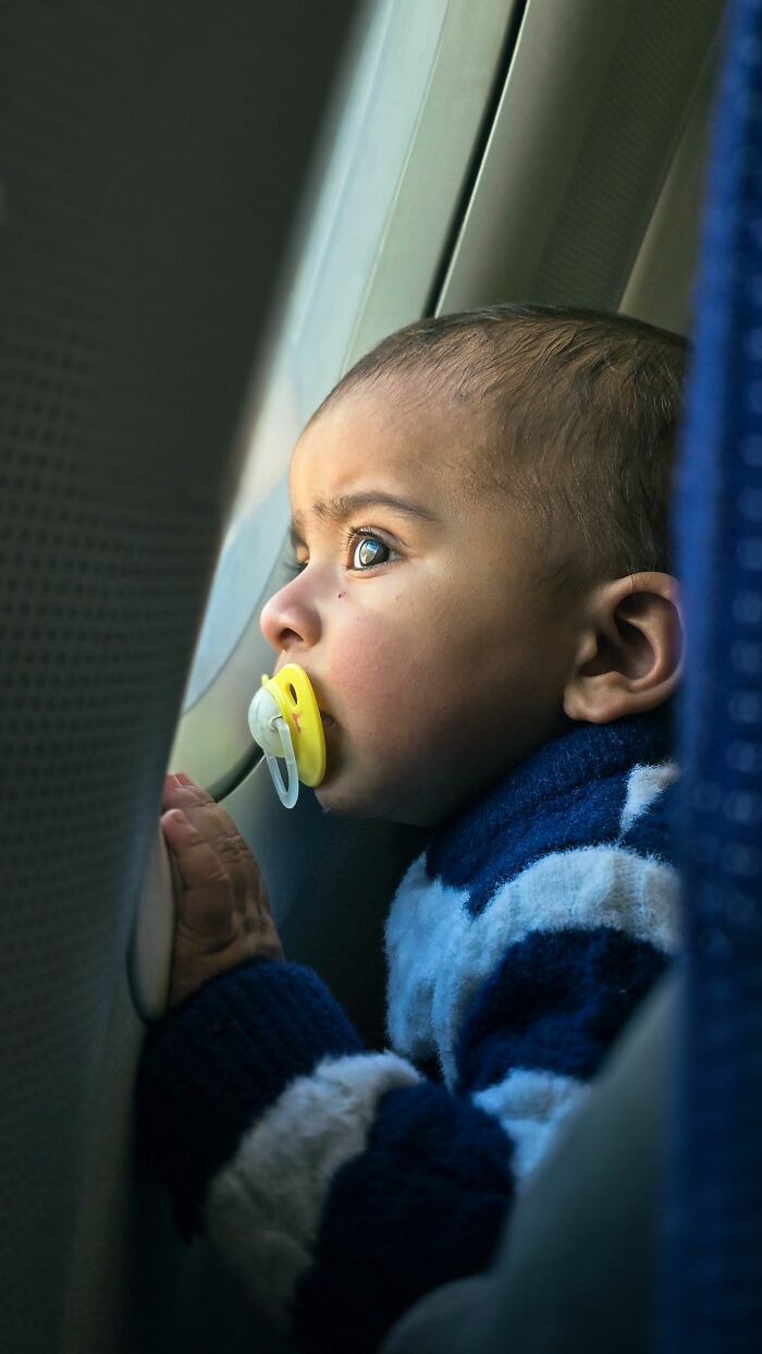 A baby with a pacifier looking out a plane window, witnessing the reality check of entitled people.