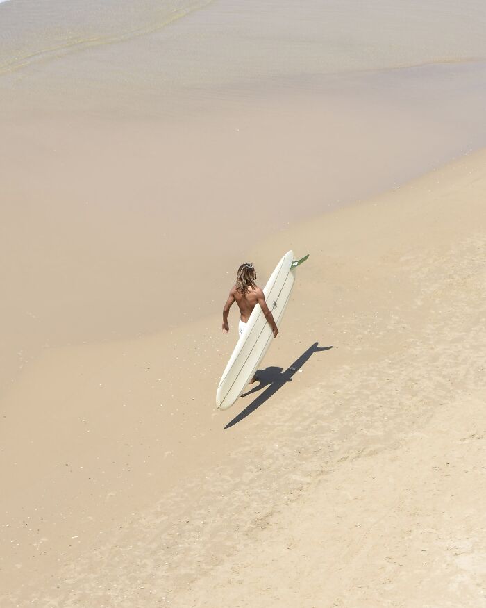 A photographer uses negative space to capture a person carrying a surfboard on a wide, minimalist sandy beach.
