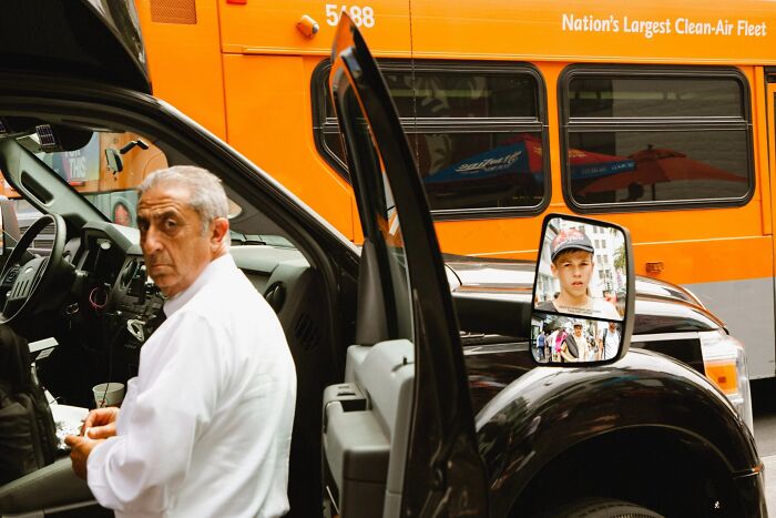 A man looks at the camera while a boy is reflected in a car mirror, a perfectly timed street photo by Jeremy Paige.
