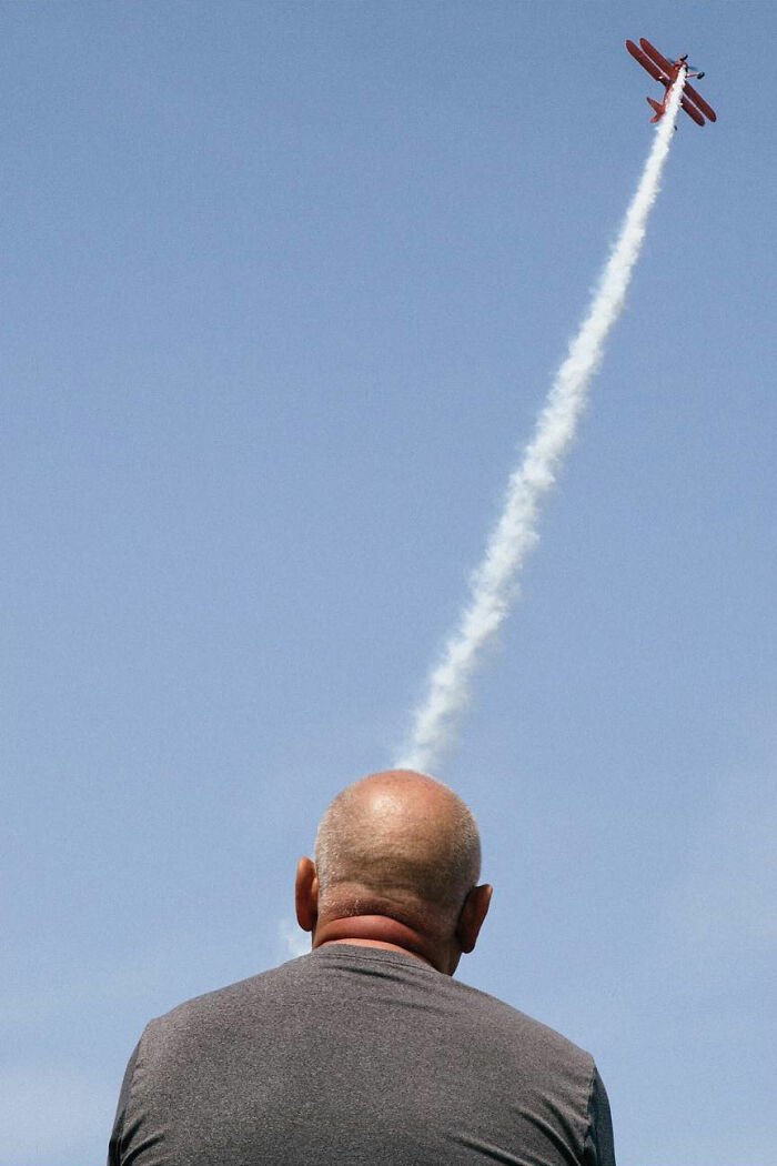 A perfectly timed street photo by Jeremy Paige of a man watching a red biplane ascend into the blue sky.