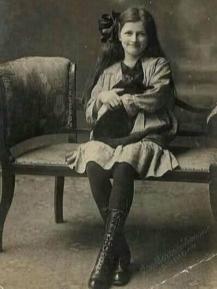 A young girl in a vintage dress sitting on a chair holding her black cat in charming vintage cat photos.