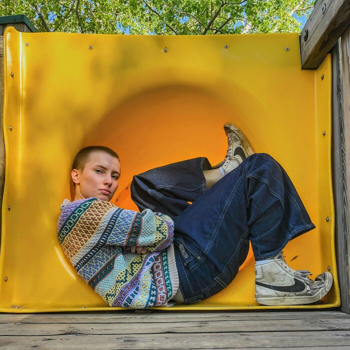 Person wearing a multicolored crocheted sweater posing in a yellow playground tunnel