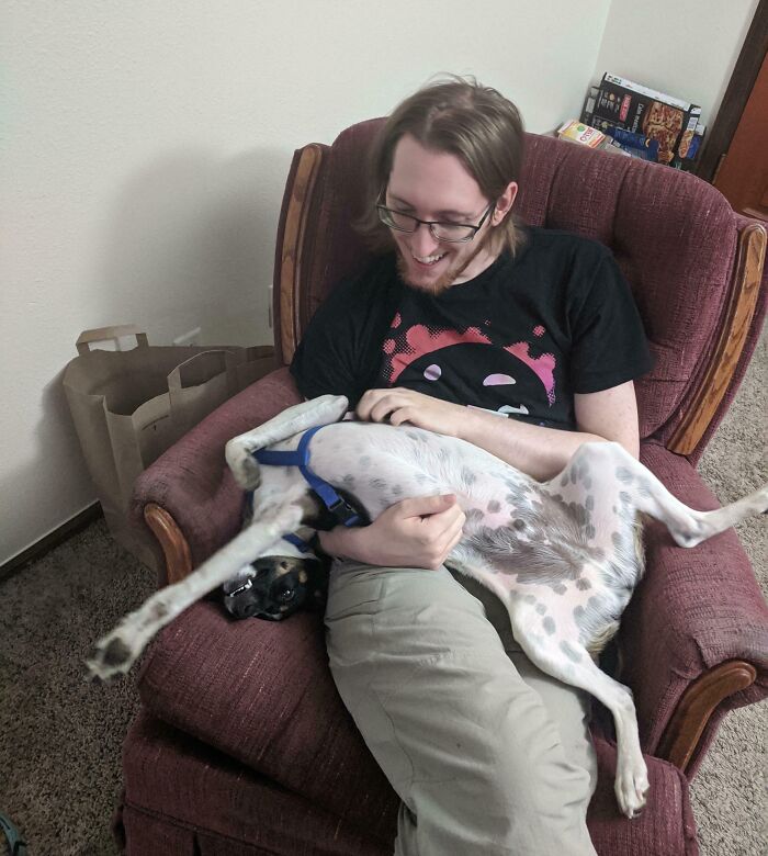 A man sitting in an armchair holding a derpy animal, a dog with white fur and black spots, looking ridiculous.