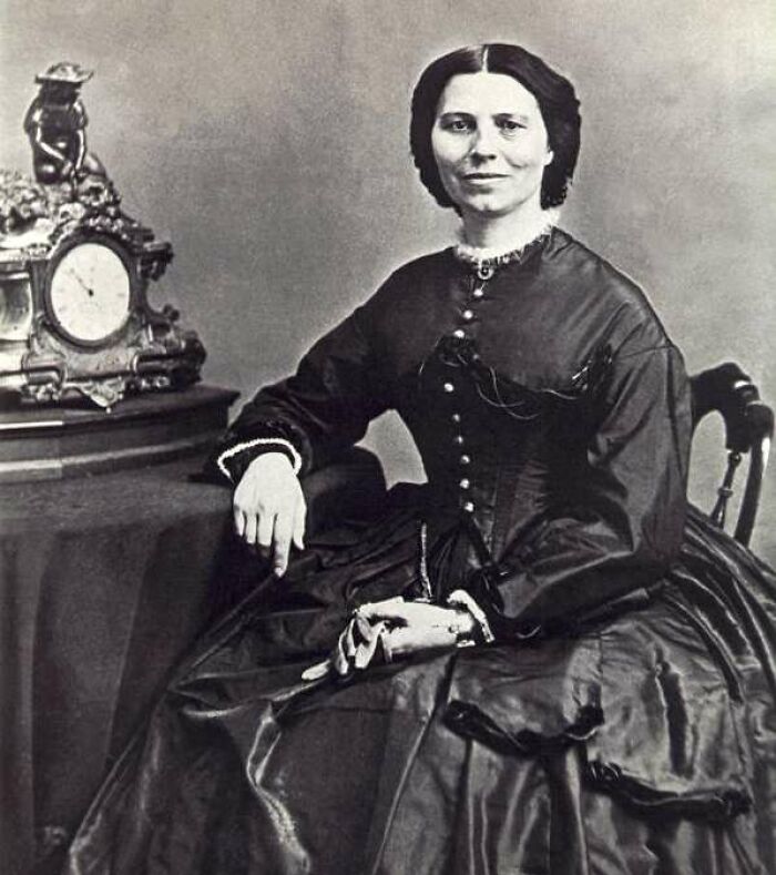 A black and white portrait of Clara Barton, a significant Women In History, seated beside an ornate clock.
