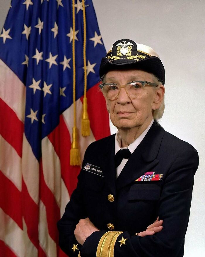 An older woman in a US Navy uniform with glasses, standing in front of an American flag, representing women in history.