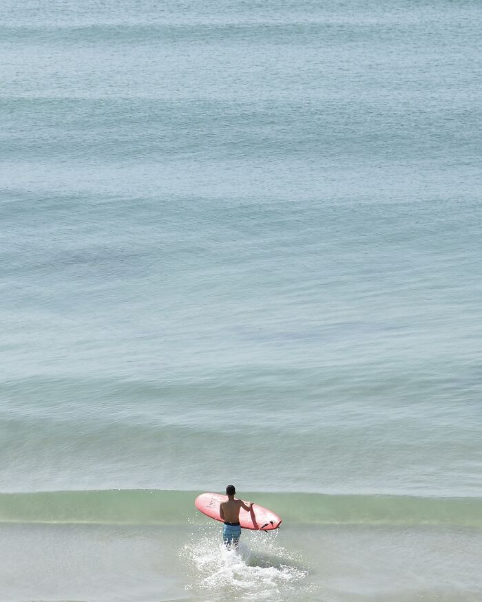 A photographer uses negative space to capture a person with a red surfboard walking into the ocean at a beach.