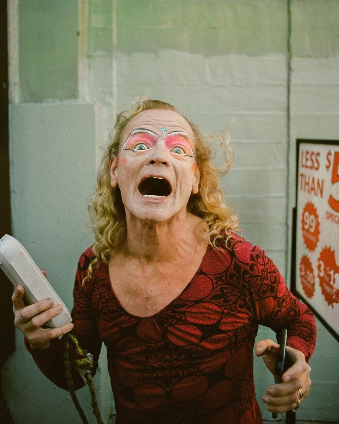 A person with dramatic makeup and an open mouth holds a phone in a perfectly timed street photo by Jeremy Paige.