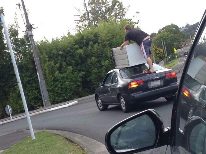 Man precariously balancing large appliance on car roof in moving fails