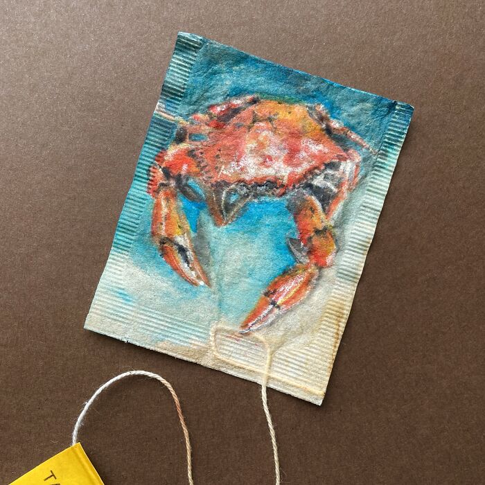 A detailed miniature painting of an orange crab on a used tea bag.