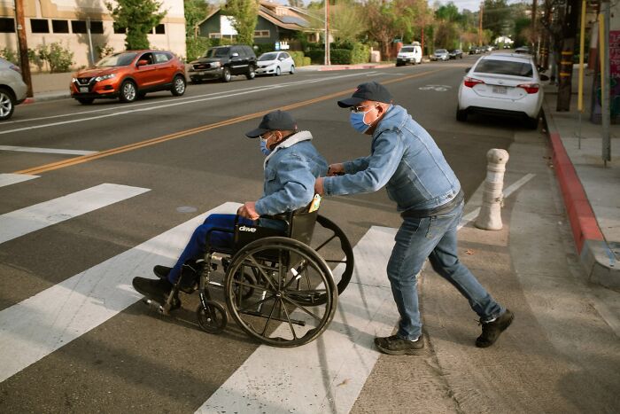 A perfectly timed street photo by Jeremy Paige of a man pushing a person in a wheelchair across a crosswalk.